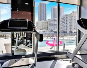 Un gimnasio con cintas de correr y equipo de ejercicio, ventanas amplias con vistas a edificios altos y una zona de piscina.