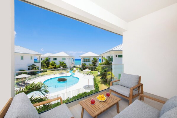 A balcony with a view of a swimming pool and resort buildings, wooden furniture, and a table with a fruit bowl and drinks.
