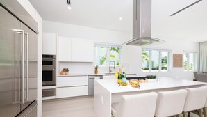 A modern kitchen with white cabinetry, stainless steel appliances, a central island with bar stools, and large windows offering a view of greenery.