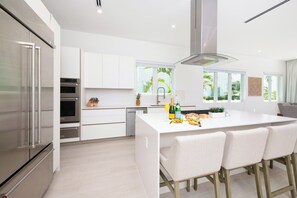 A modern kitchen with white cabinetry, stainless steel appliances, a central island with bar stools, and large windows offering a view of greenery. - Oceans 9, Villa 8: Lazy River Pool, Sunken BBQ Lounge + Designer Interiors in West Bay (Grand Cayman)