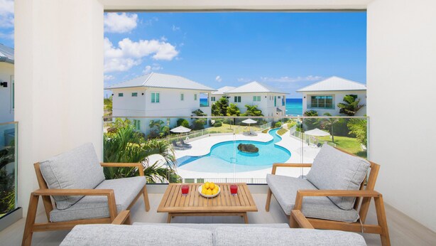 A balcony with a view of a swimming pool and houses, wooden furniture, and a table with fruit.