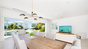 A modern living room with a wooden dining table, grey upholstered chairs, a large flat-screen TV, and a ceiling fan.