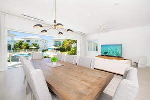 A modern living room with a wooden dining table, grey upholstered chairs, a large flat-screen TV, and a ceiling fan. - Oceans 9, Villa 8: Lazy River Pool, Sunken BBQ Lounge + Designer Interiors in West Bay (Grand Cayman)