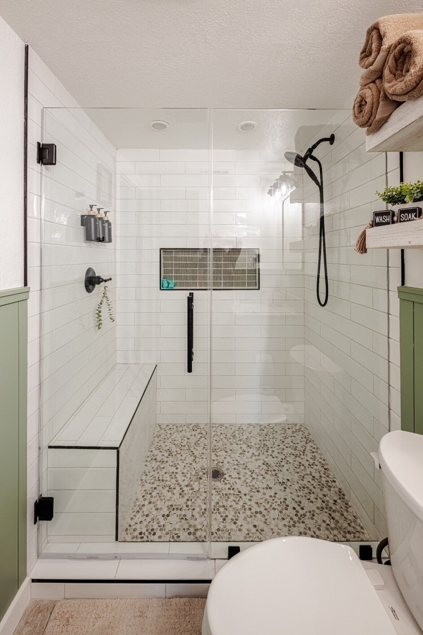 A glass-enclosed shower with a white tiled floor, a toilet, and a shelf with towels.