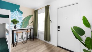 Une pièce comprenant un lit superposé, un petit bureau avec des tabourets, une plante en pot, une porte et une fenêtre avec des rideaux.
