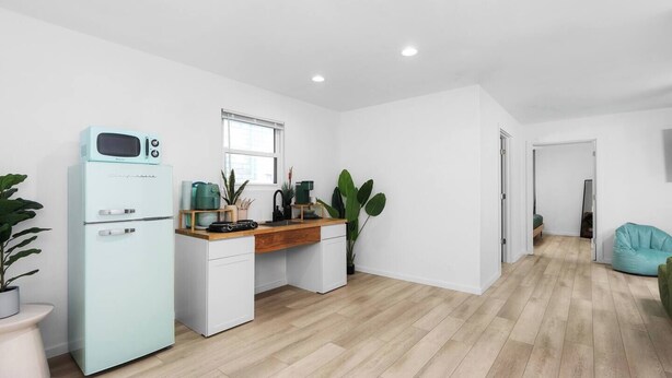 Une cuisine moderne équipée d’un four à micro-ondes encastré, d’un évier et d’une fenêtre. On y trouve un bureau en bois, une plante et une machine à café. La pièce est dotée d’un plancher en bois et de murs blancs.