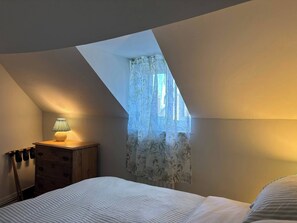 A bedroom with a sloped ceiling, a bed with striped bedding, a bedside table with a lamp, a wooden dresser, and a window with floral curtains. - Quaint Townhouse in the Centre of Chipping Norton (Chipping Norton)