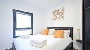 A neatly made bed with white linens and orange pillows, a window with blinds, and a wall-mounted abstract art piece. - Hayarkon Joie in Tel Aviv-yafo (Tel Aviv)