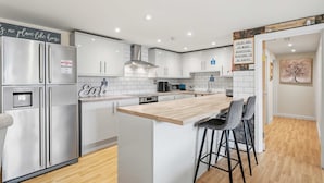 A modern kitchen with stainless steel appliances, white cabinetry, and a wooden island with bar stools. - 6-bedroom Family Haven With Garden in Aylesbury (Aylesbury)