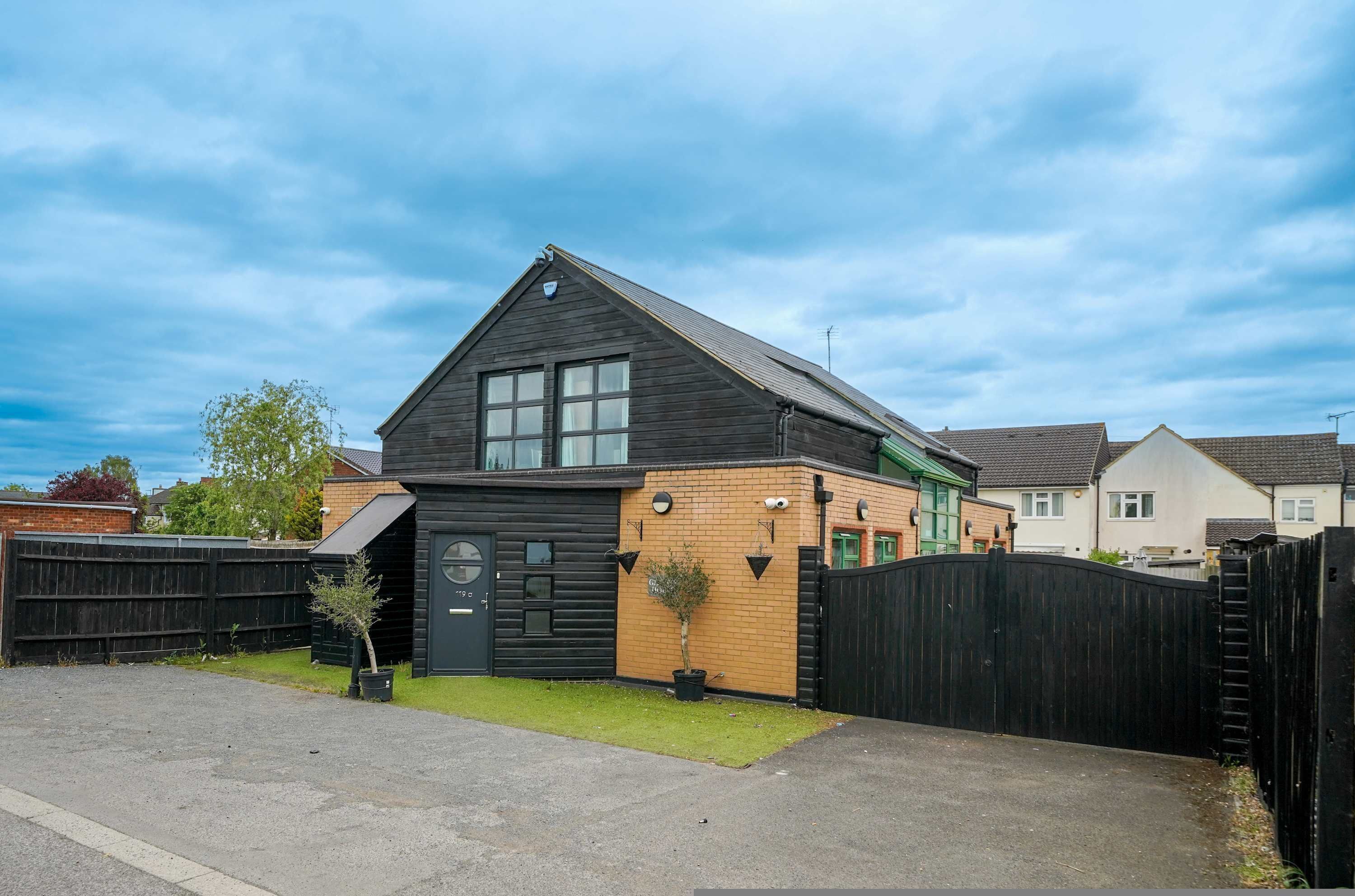 A residential house with a dark roof, a brick extension, a black gate, and a small tree in the yard.