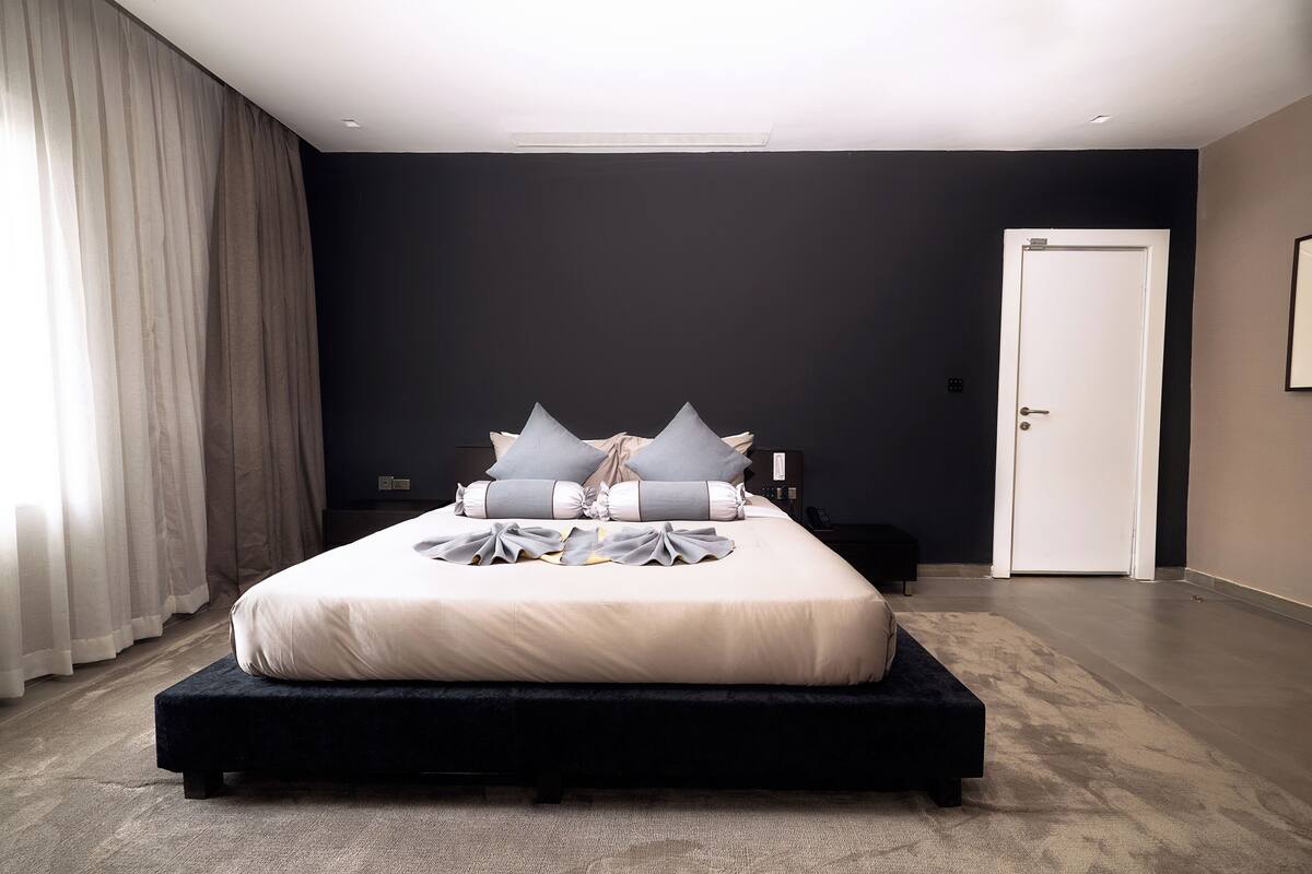 A modern bedroom with a large bed, a dark wall, a white door, and a large window with curtains.