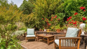 A wooden deck with two chairs and a table, surrounded by lush greenery and flowering plants. - Coquelicot Guest Room with Shared Pool, Wi-Fi, and Air Conditioning (Lagarde-Paréol)