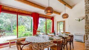 A dining area with a round table, chairs, a patterned tablecloth, red curtains, and a view of trees through large windows. - Coquelicot Guest Room with Shared Pool, Wi-Fi, and Air Conditioning (Lagarde-Paréol)