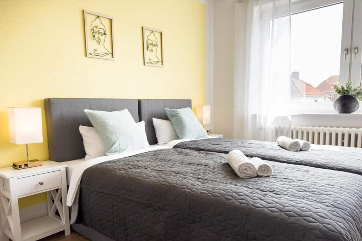 Ein Schlafzimmer mit einem Bett, Nachttischen, einem Fenster und zwei an der Wand befestigten Kunstwerken.