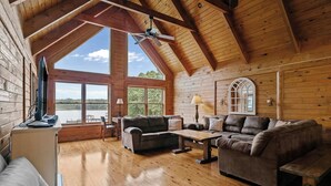 Ein Blockhaus-Interieur mit Holzbalkendecke, großen Fenstern, einer Couch, einem Tisch und einem Fernseher.