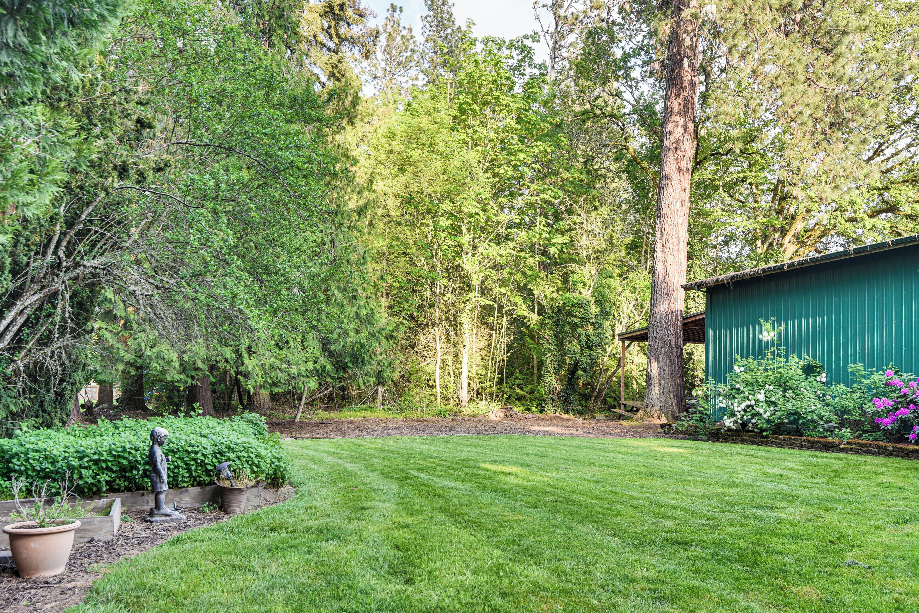 Une cour bien entretenue, recouverte de gazon, avec une terrasse en bois, entourée d’arbres touffus et d’un abri de jardin vert.