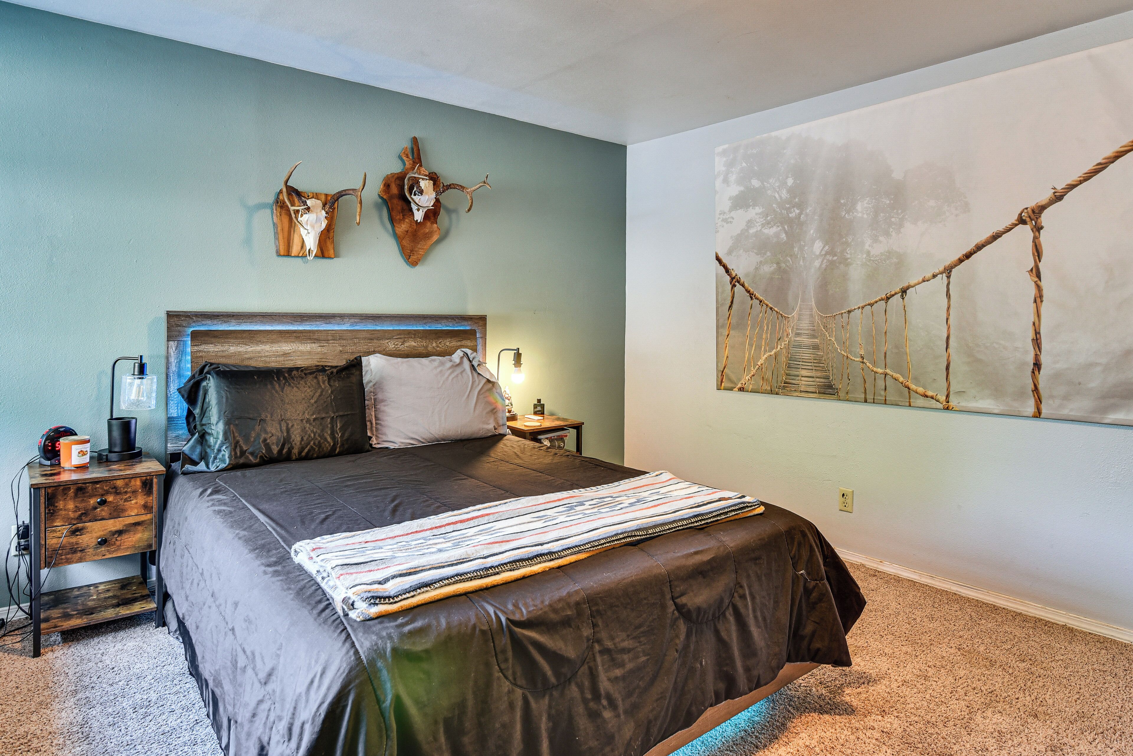 A bedroom with a bed, wooden headboard, two wall-mounted animal skulls, a rope bridge painting, and a nightstand with a lamp.