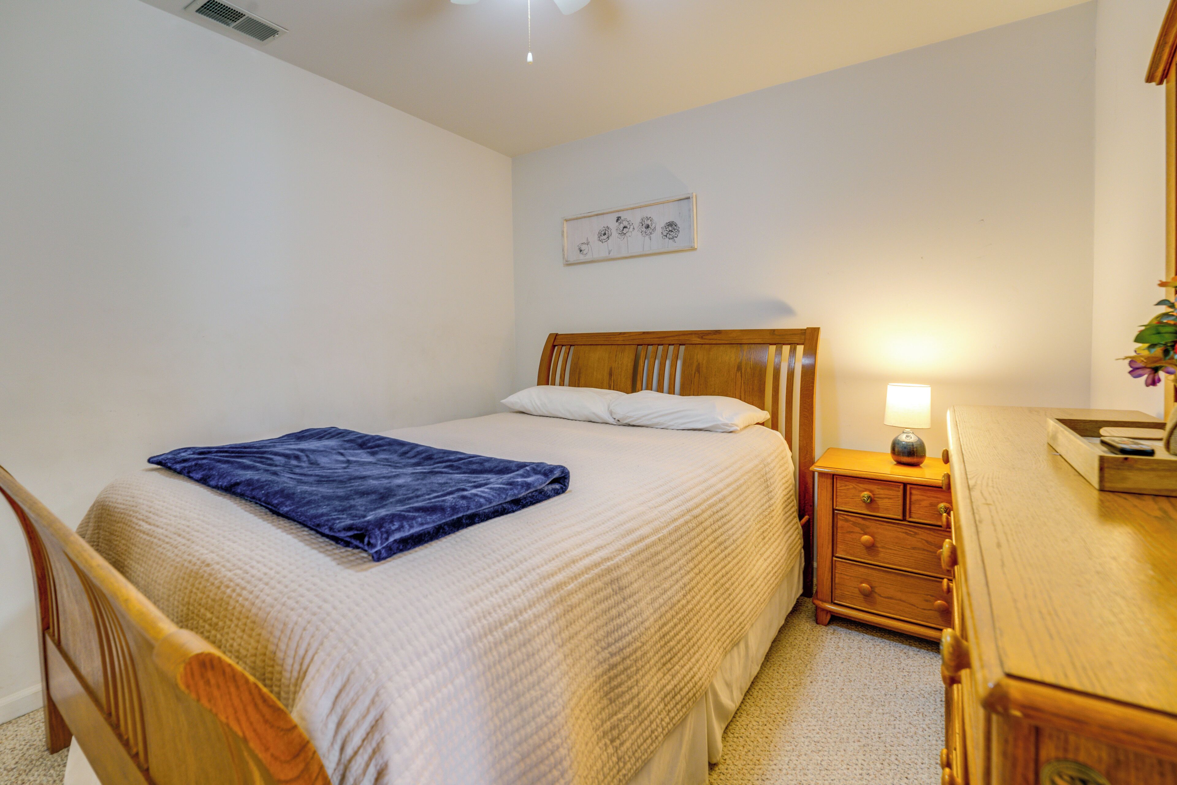 A bedroom with a bed, a wooden nightstand with a lamp, a dresser, and a wall-mounted picture.