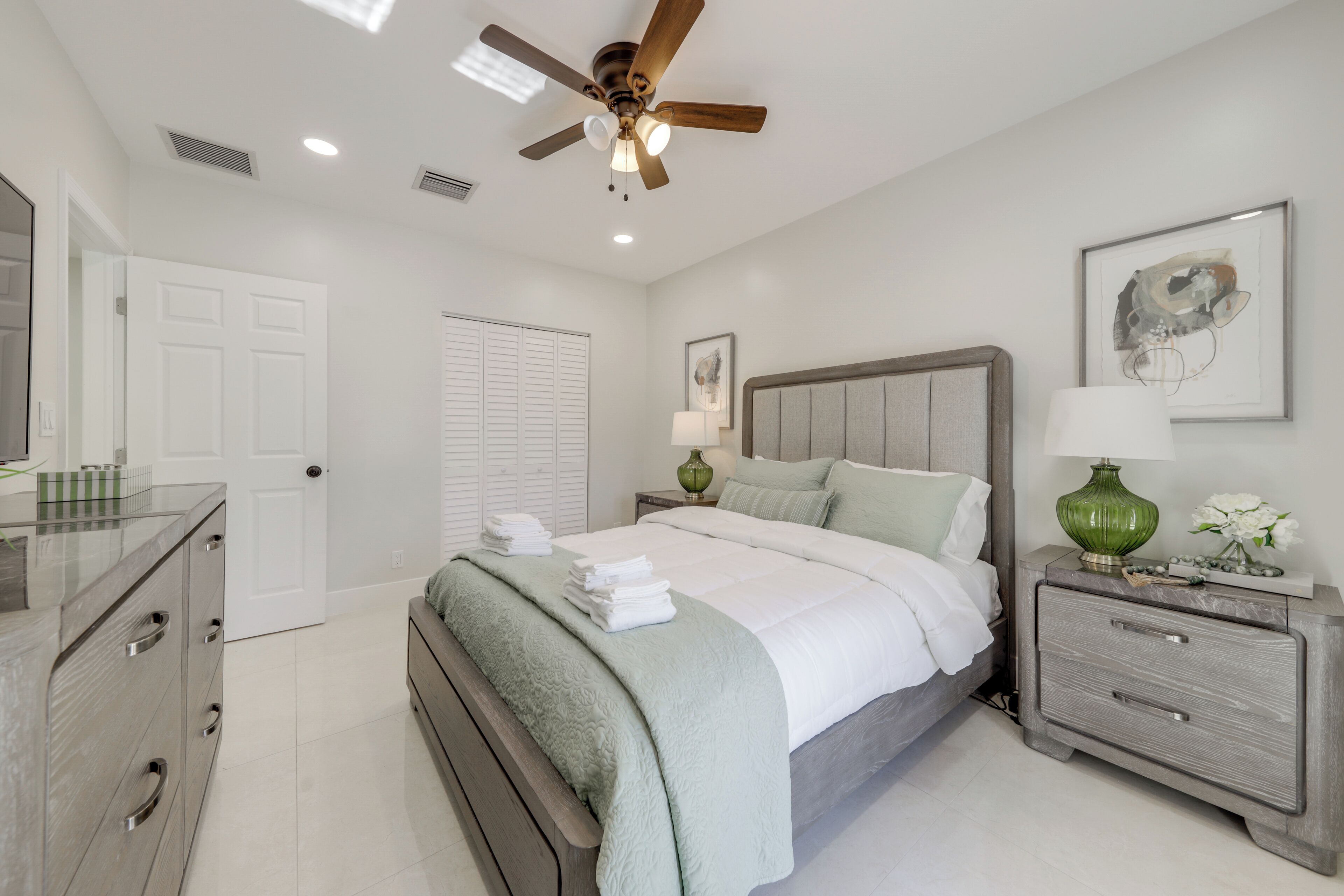 Une chambre à coucher comprenant un lit, des tables de chevet, une commode, un ventilateur de plafond et une porte.