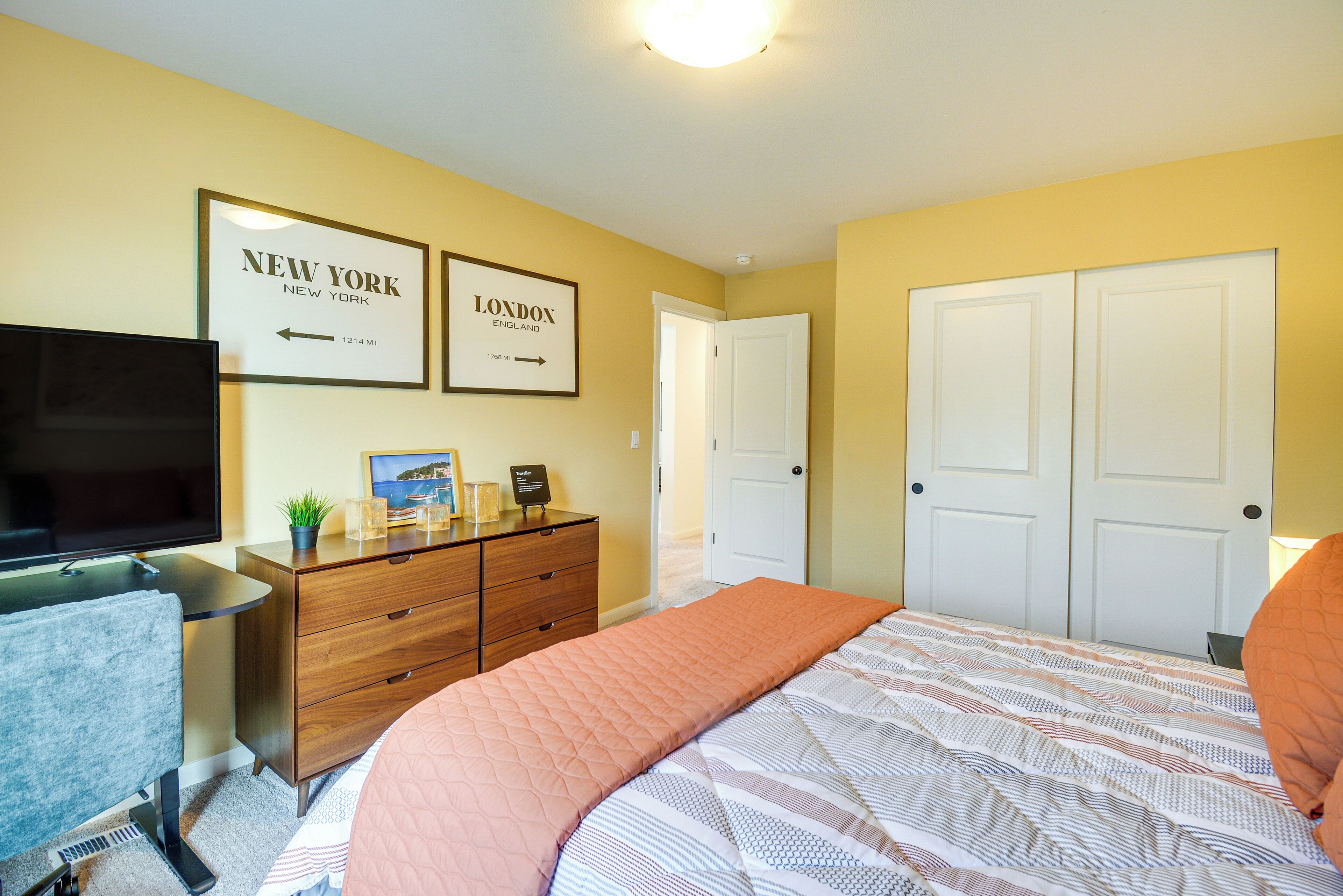 A hotel room with a bed, a desk with a TV, a dresser, and two doors.