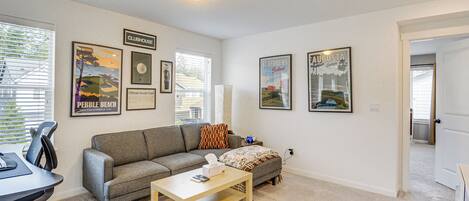 Una sala de estar con un sofá gris, una mesa de centro de madera y un televisor. Hay cuadros enmarcados en la pared, una ventana con persianas y una puerta que conduce a otra habitación.