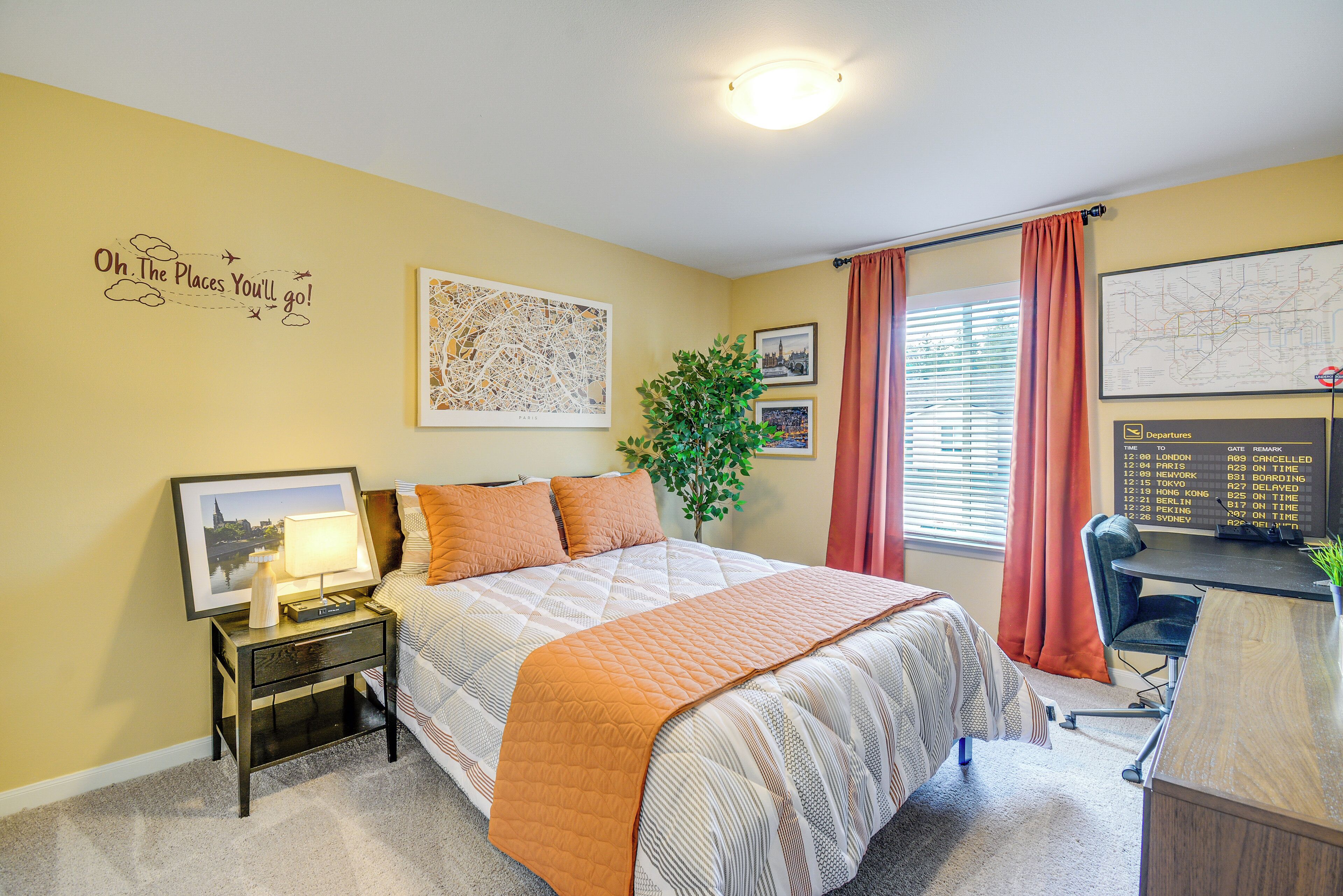 A hotel room with a bed, a desk, a nightstand, a map on the wall, and a plant.