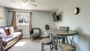 A living room with a sofa, a chair, a round table with stools, a ceiling fan, a window with curtains, and a wall-mounted TV. - Explore Port Huron! Cozy Condo w/ Creek Views (Port Huron)
