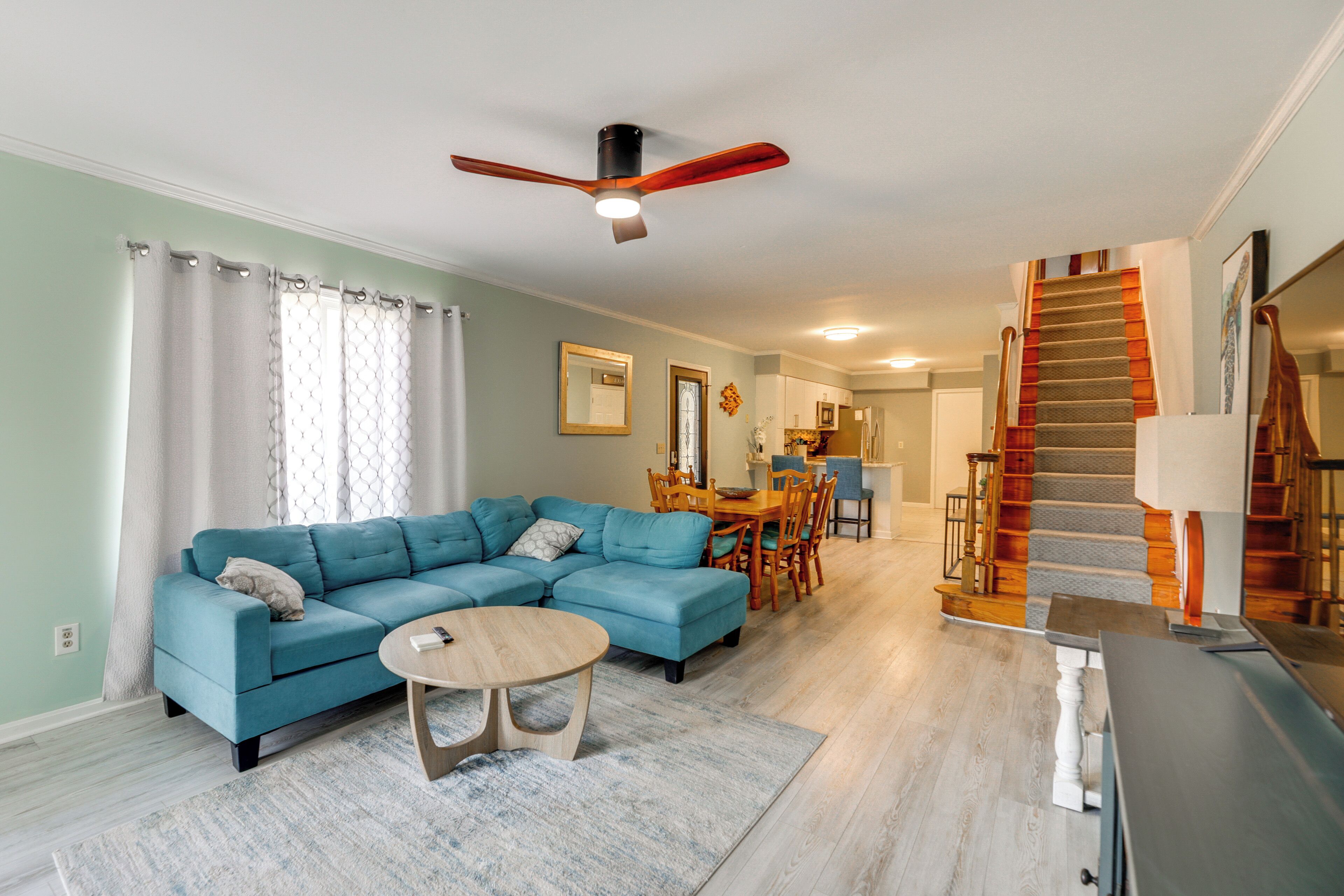 A living room with a sectional sofa, a dining table with chairs, a ceiling fan, and a staircase.