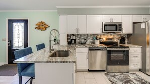 A modern kitchen with white cabinets, stainless steel appliances, a marble countertop, and a dining area with blue chairs. - Easy Beach Access! Bethany Retreat on Trolley Loop (Bethany Beach)