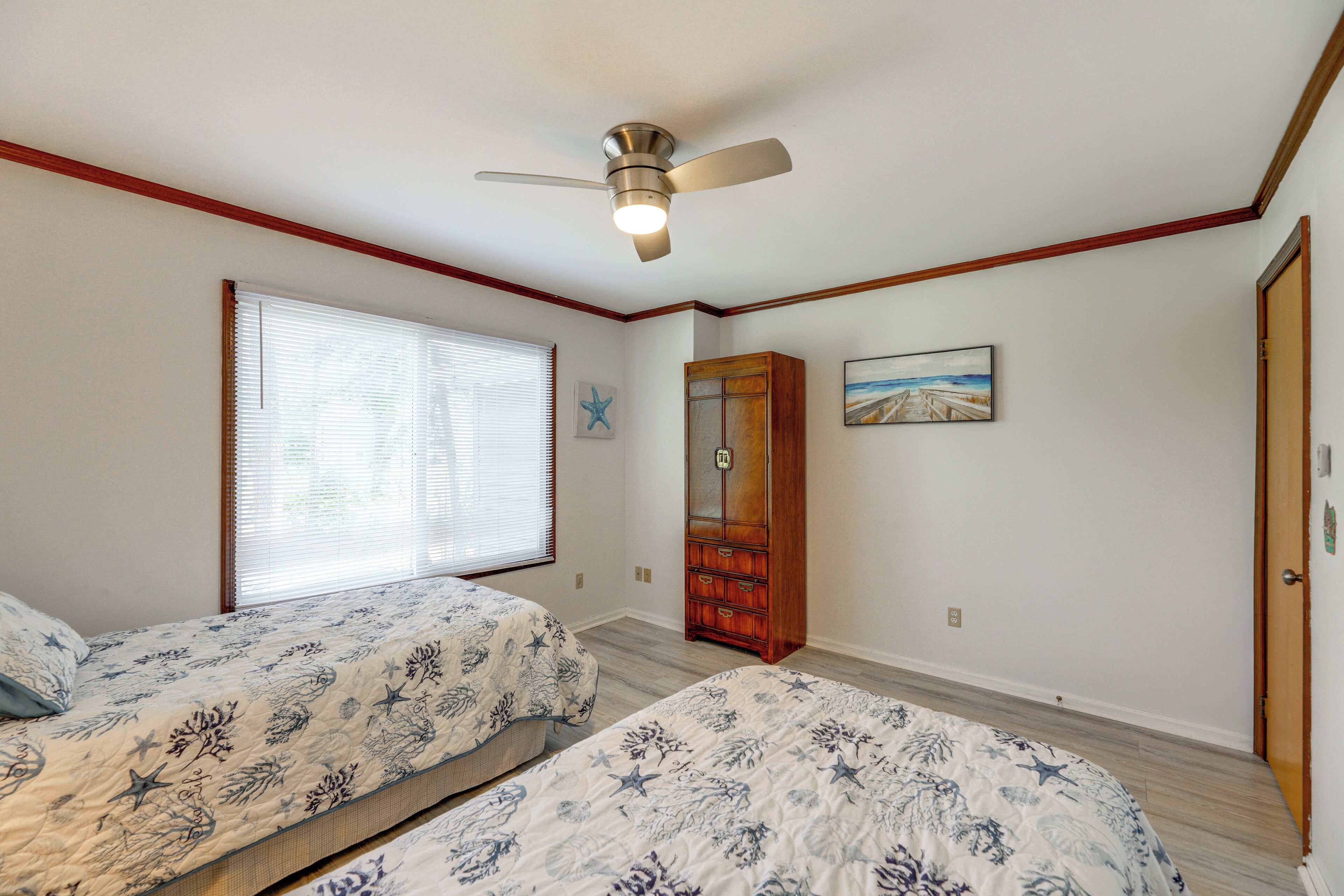 Une chambre avec deux lits, un ventilateur de plafond, une fenêtre avec des stores, une armoire en bois et un tableau au mur.