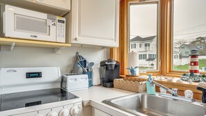 A kitchen with a stove, microwave, and sink, with a view of houses and a lighthouse through the window. - Coastal Hampton Condo: Walk to North Beach! (Hampton)