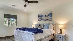 Une chambre à coucher avec un lit, des tables de chevet, une lampe, un ventilateur de plafond, un tableau et une fenêtre.
