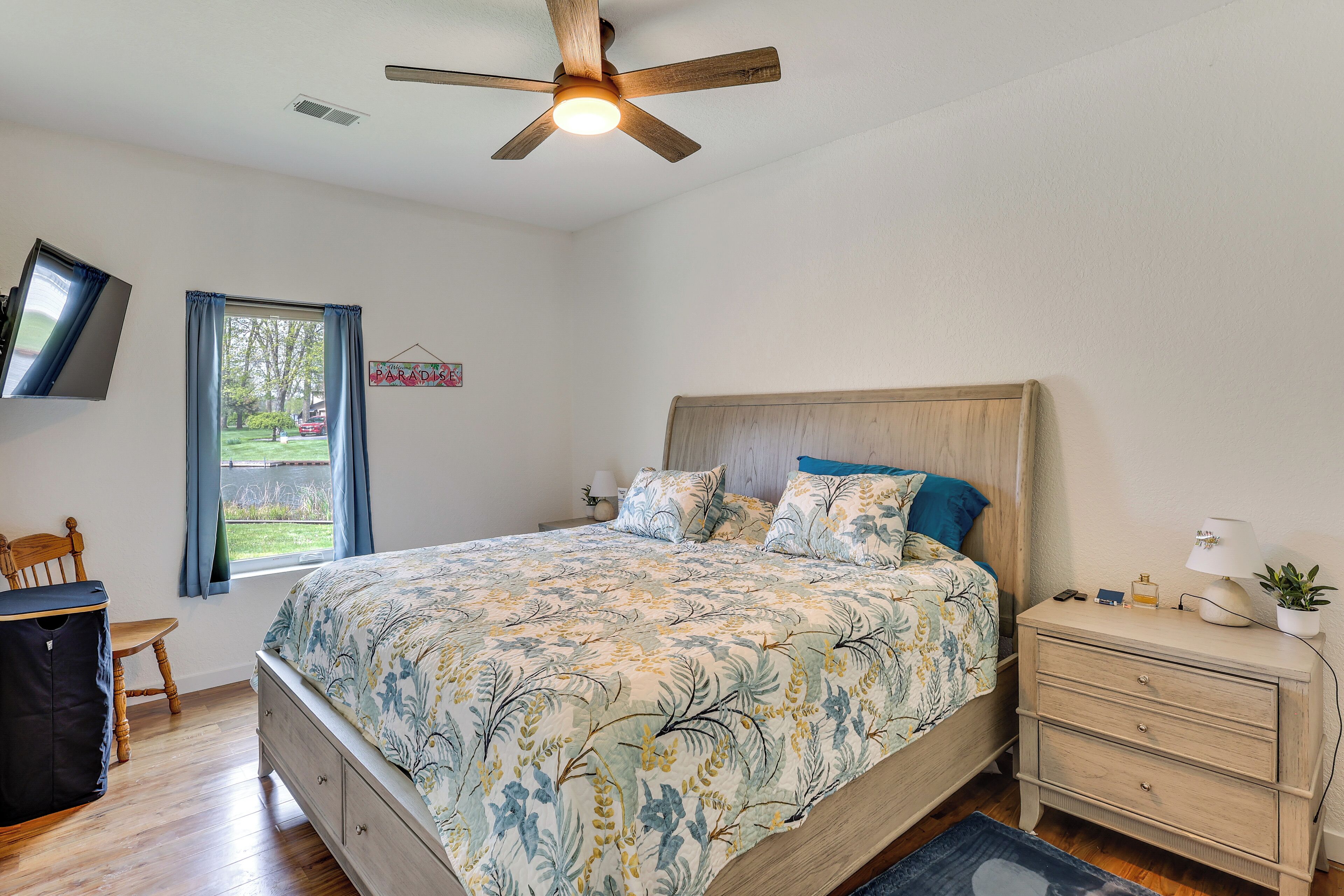A bedroom with a bed, a nightstand, a ceiling fan, a window with curtains, a chair, and a TV mounted on the wall.