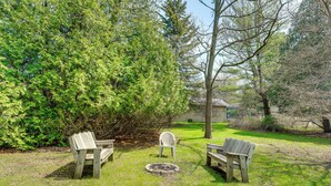 Un espace extérieur verdoyant avec deux bancs en bois et une chaise en plastique blanche, entouré d’arbres et avec un bâtiment en arrière-plan.