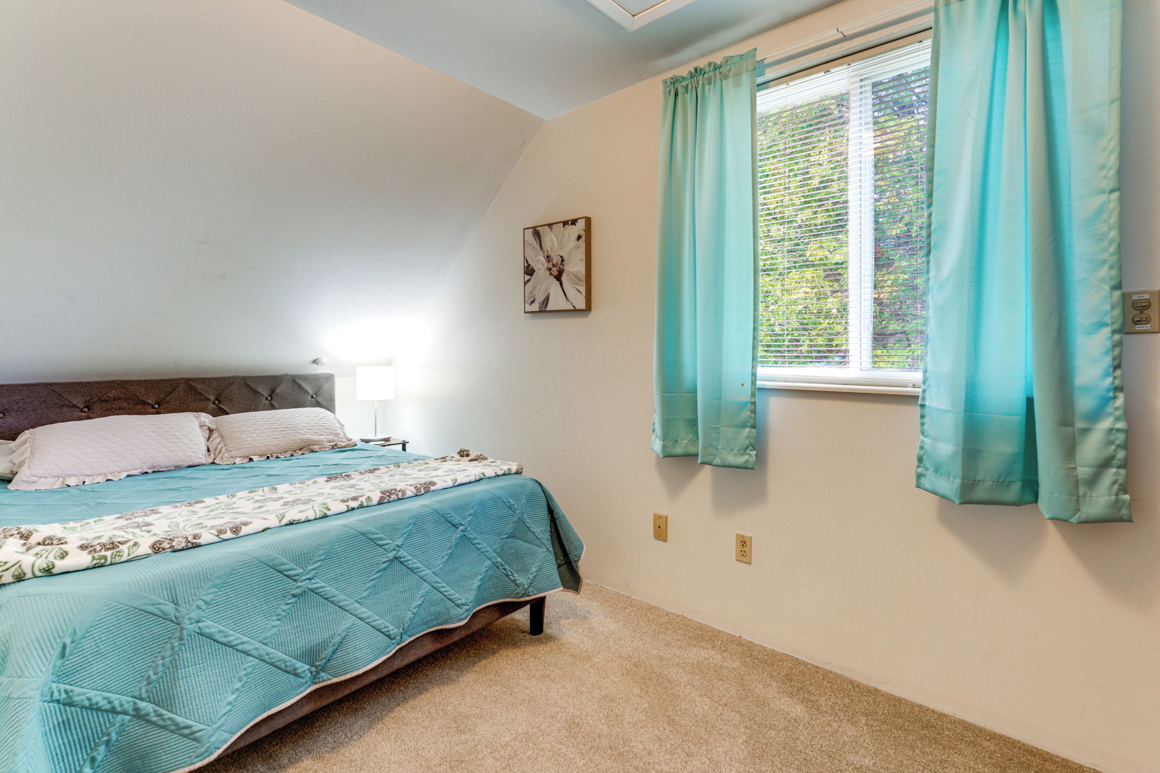 A bedroom with a bed, a window with curtains, a wall-mounted picture, and a carpeted floor.