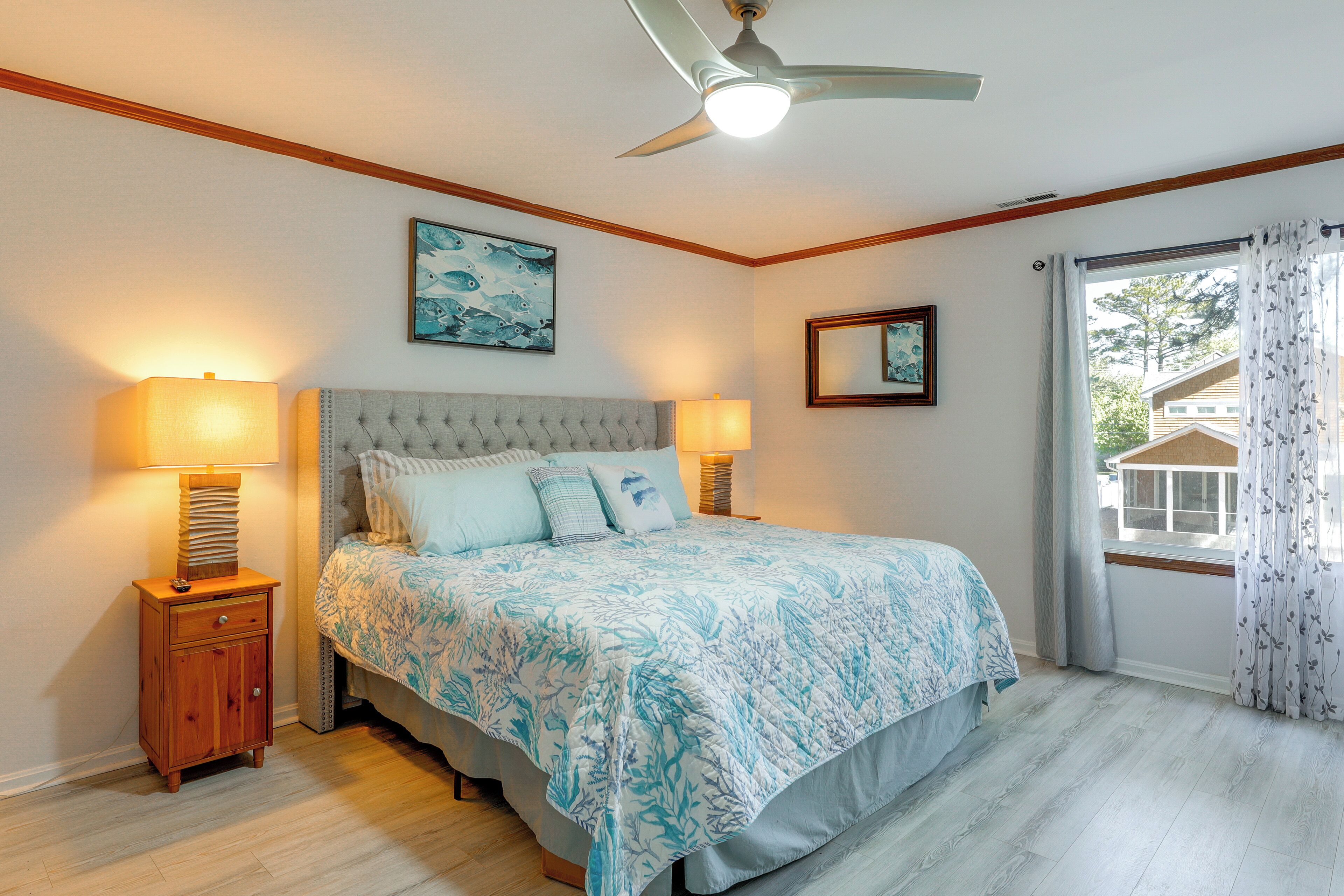 Une chambre à coucher comprenant un lit, deux lampes de chevet, un ventilateur de plafond, une fenêtre avec des rideaux et un miroir fixé au mur.