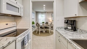 A kitchen with white cabinets, a microwave, an oven, a coffee maker, and a dining area with a table and chairs. - Pool Time & Island Vibes! Hilton Head Getaway (Hilton Head Island)