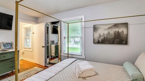 A bedroom with a bed, a bedside table, a mirror, a window with blinds, a painting on the wall, and a door leading to another room.