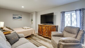 Uma sala de estar com sofá, poltrona, televisão sobre um aparador de madeira, cama com colcha xadrez e janela com cortinas estampadas.