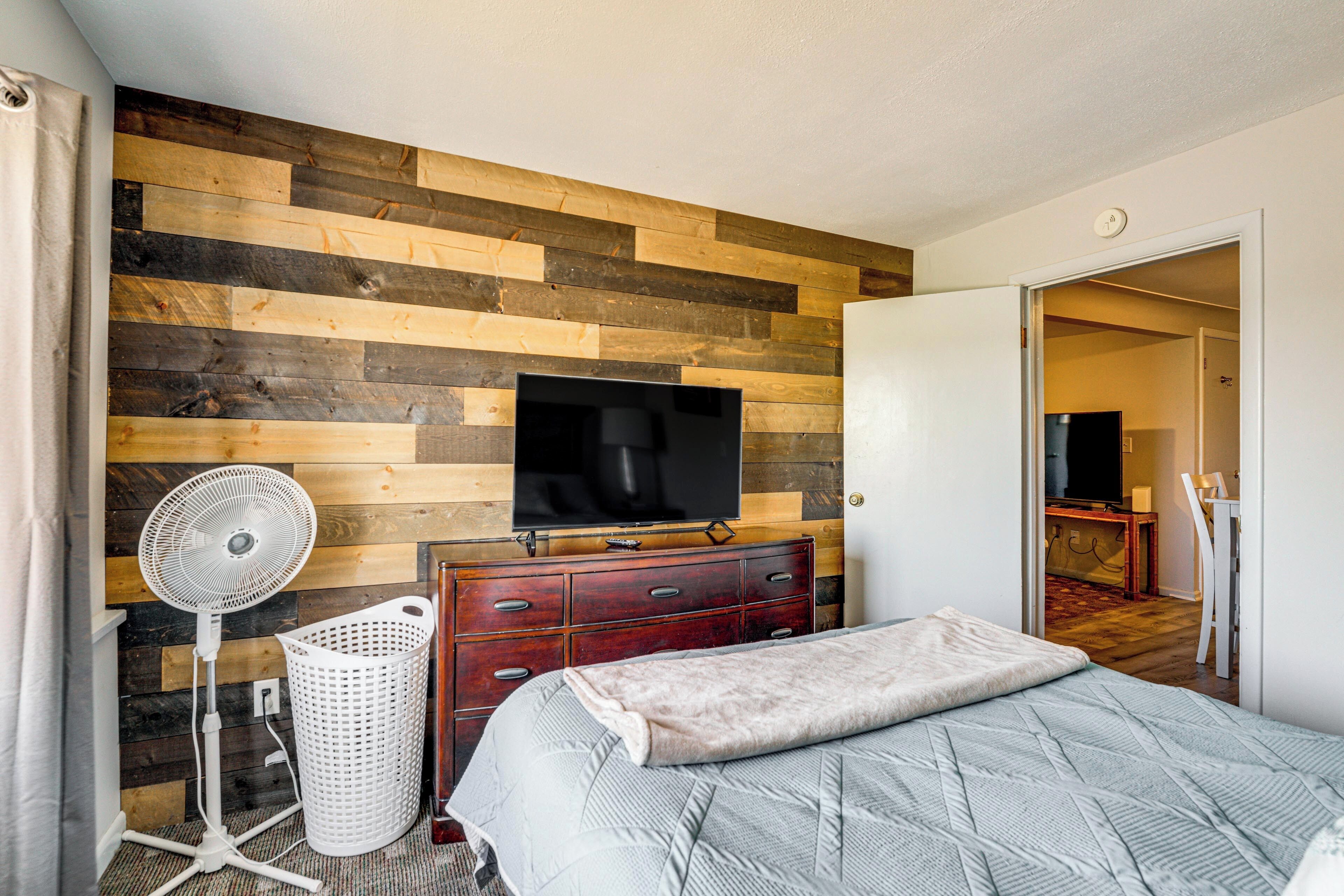 A bedroom with a wooden wall, a TV, a fan, a dresser, and a bed.