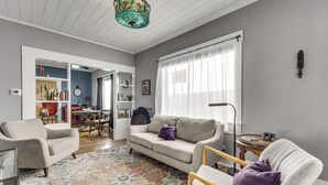 Uma sala de estar com sofá, poltronas, tapete estampado, lustre e canto de jantar com mesa e cadeiras.