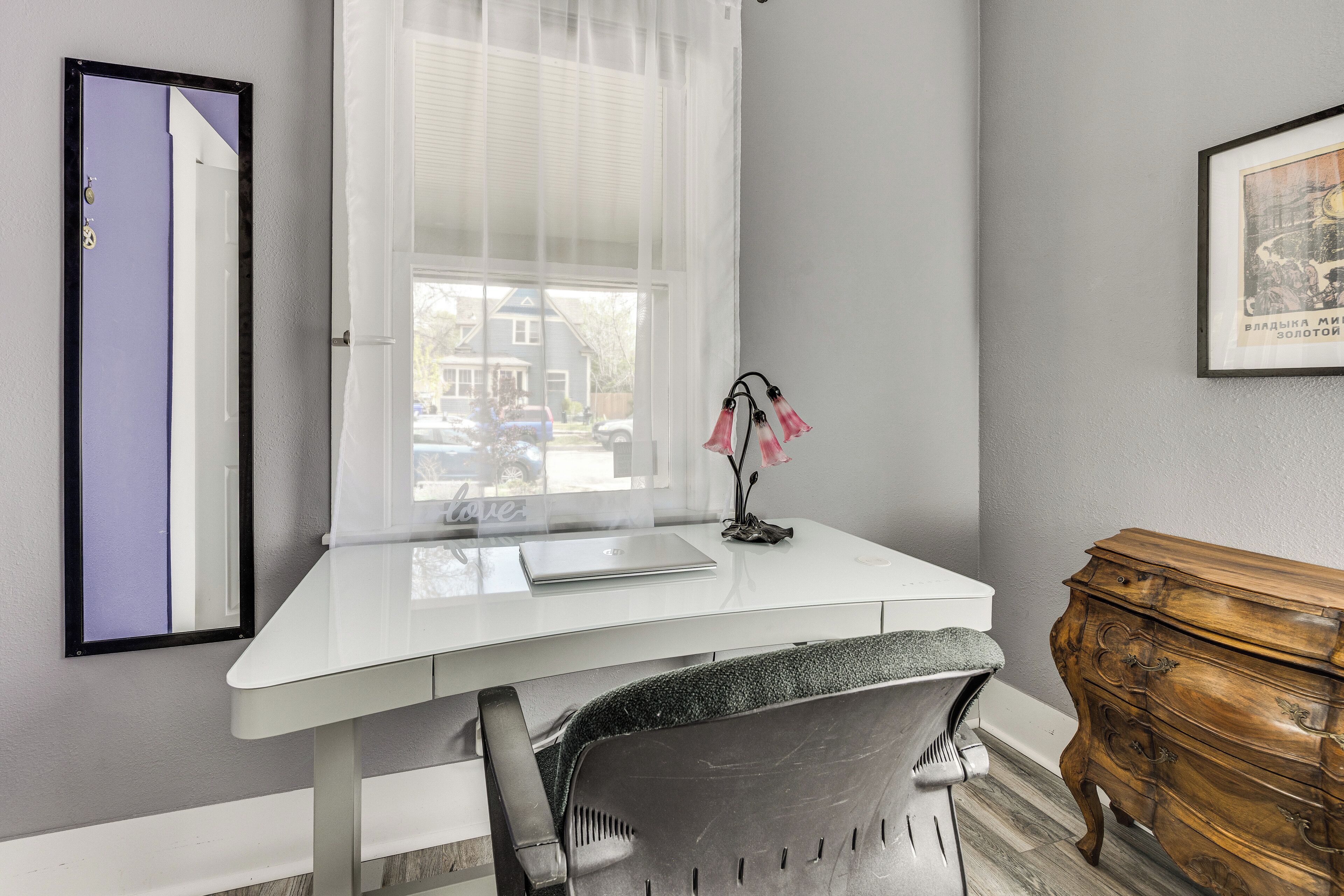 Una habitación con un escritorio blanco, una silla, un aparador de madera, un espejo y una ventana con persianas.