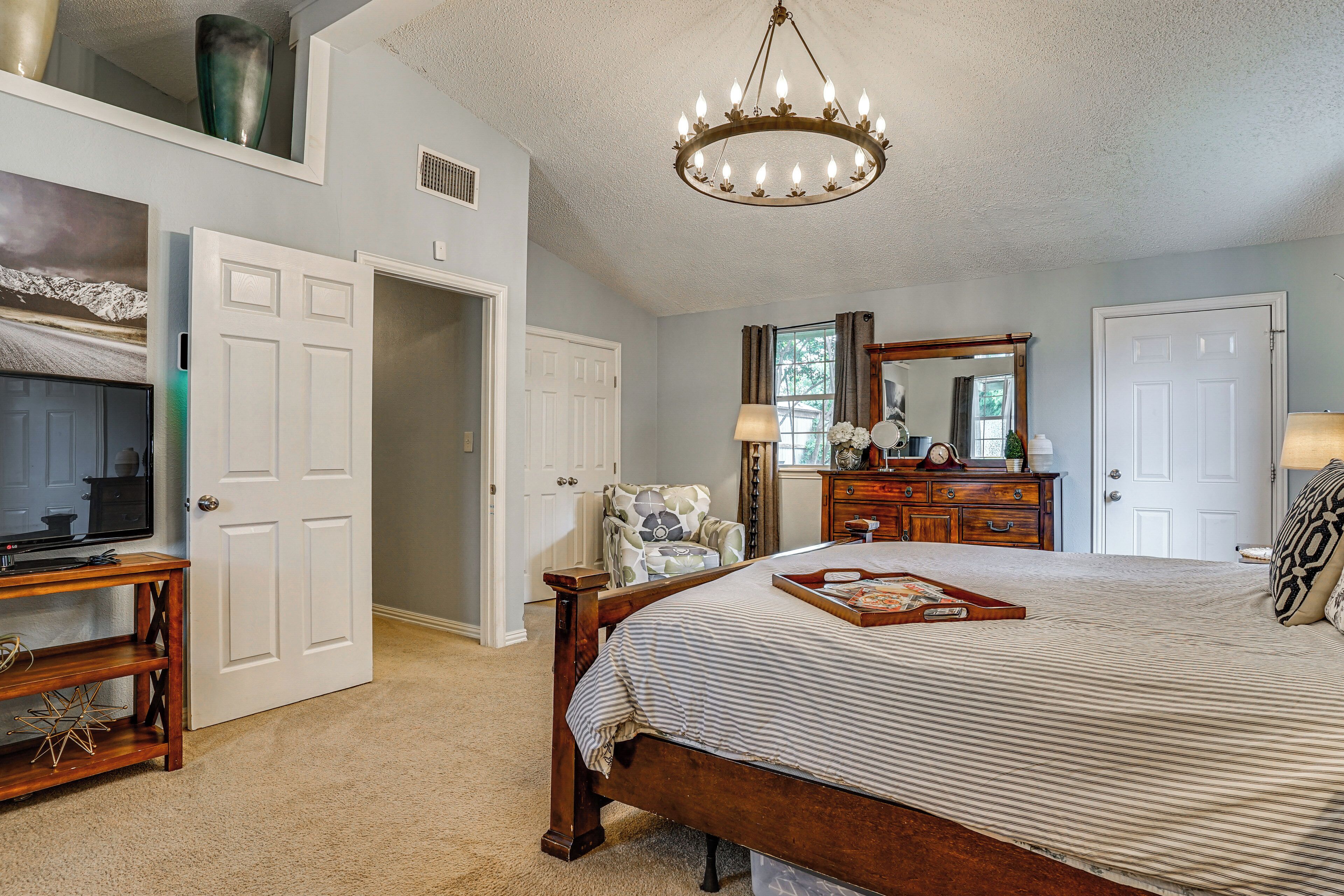A bedroom with a large bed, a television, a chandelier, a dresser, a chair, and a mirror.