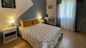 Un dormitorio con cama, mesitas de noche, un escritorio, un ventilador de techo y una ventana con cortinas.