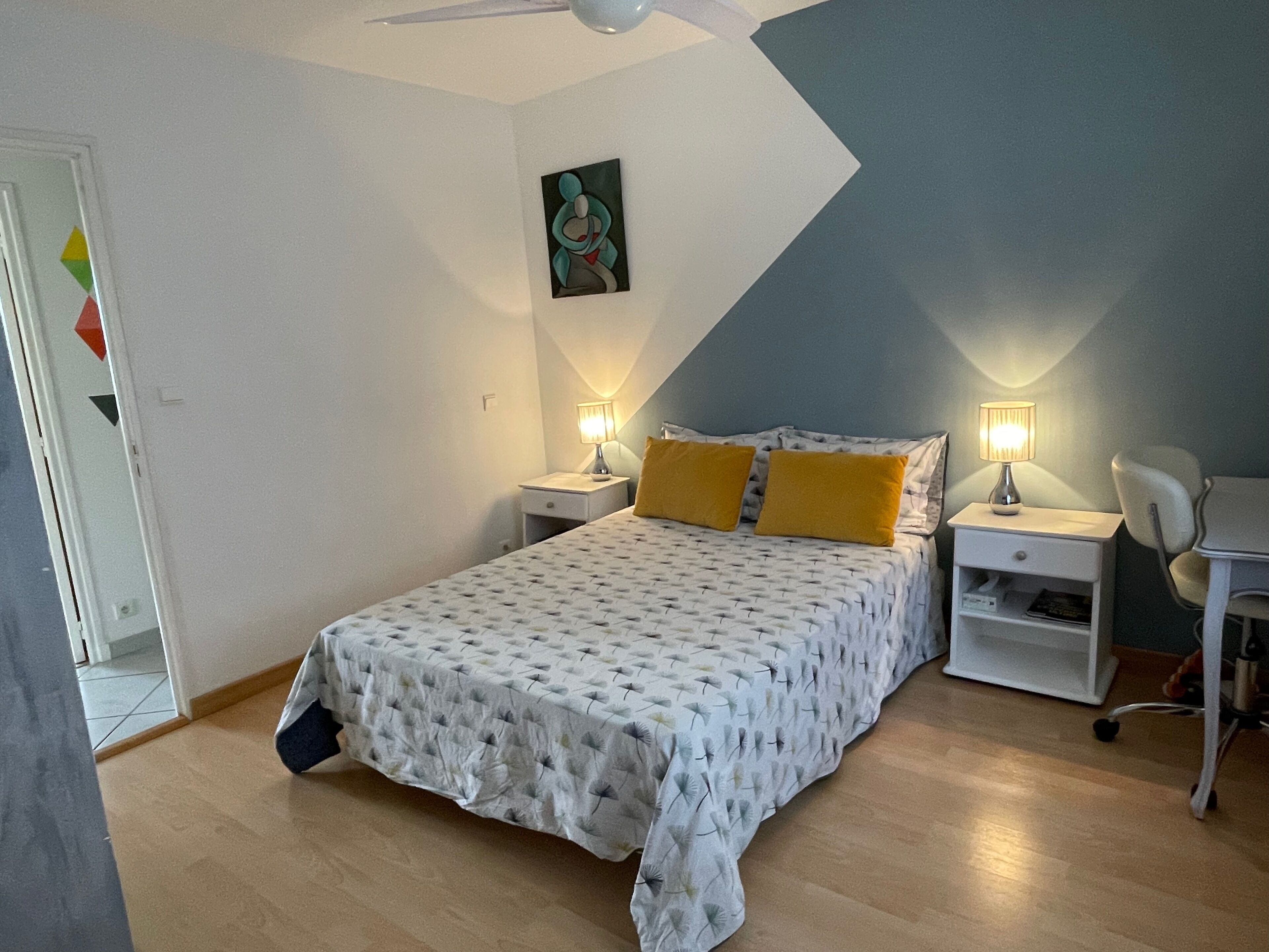 A bedroom with a bed, two bedside tables with lamps, a chair, a painting on the wall, and a door with a geometric decoration.