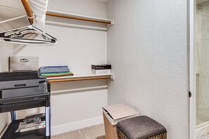 A corner of a room with a printer, a small table with a cushion, a hanging garment rack, and a shelf with folded clothes and a box. - Community Perks! Modern PHX Condo: Extended Stay! (Phoenix)
