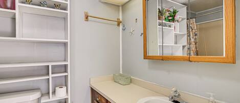 A bathroom with a toilet, sink, mirror, shelves with toiletries, and a shower curtain.
