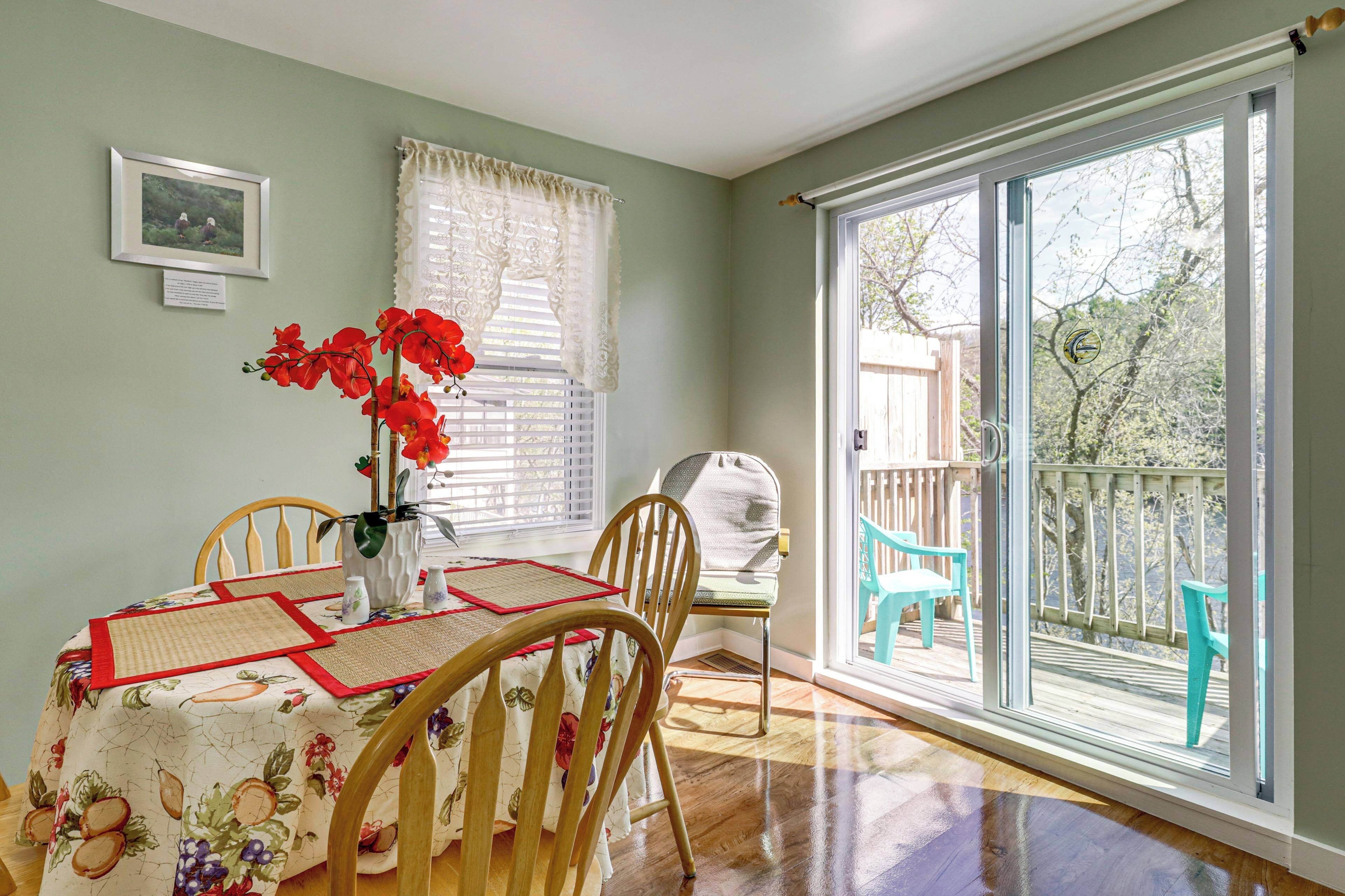 A dining area with a table set for two, a chair, a vase with red flowers, and a window with blinds.