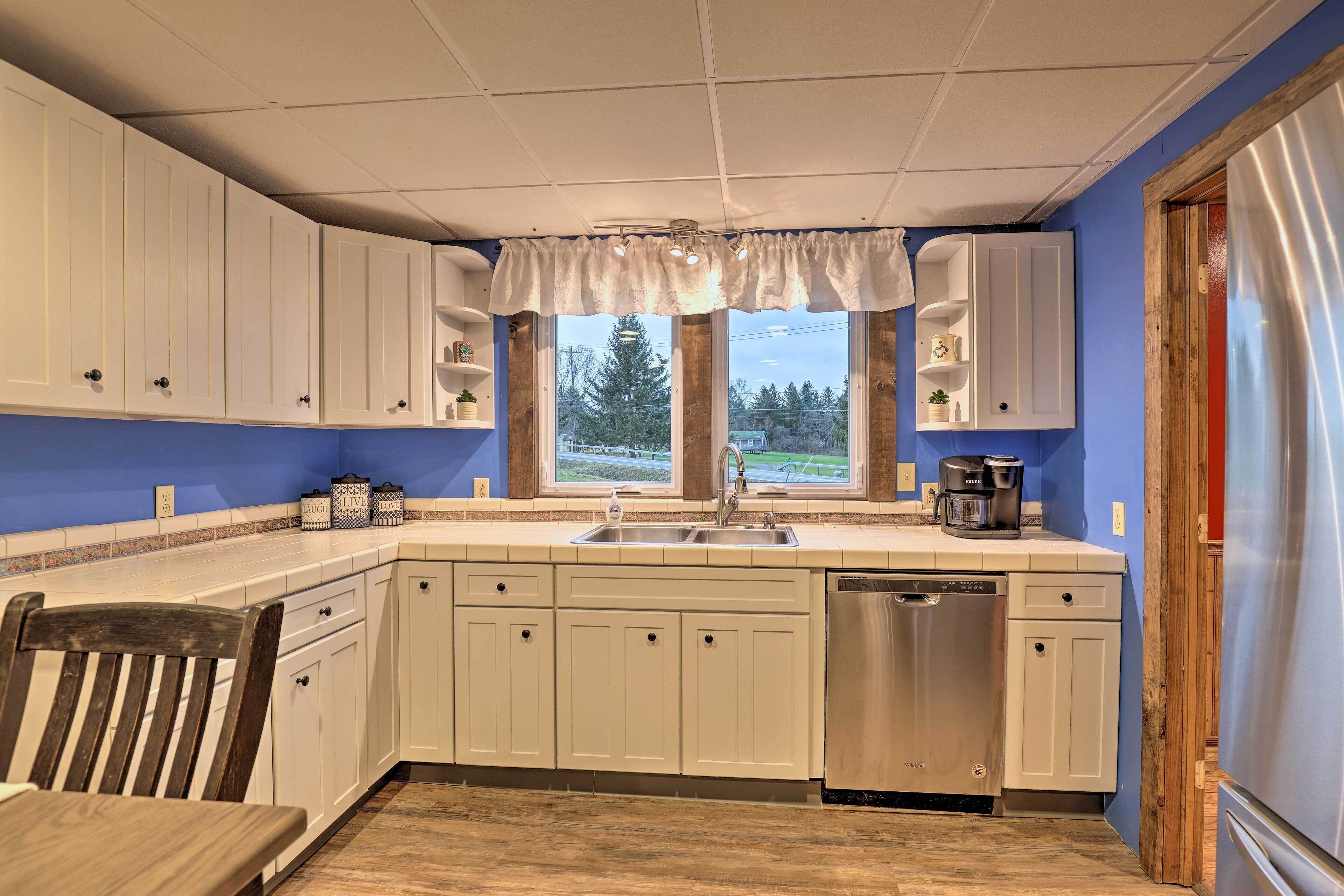 A kitchen with white cabinets, a stainless steel dishwasher, a coffee maker, and a window with a view of trees.