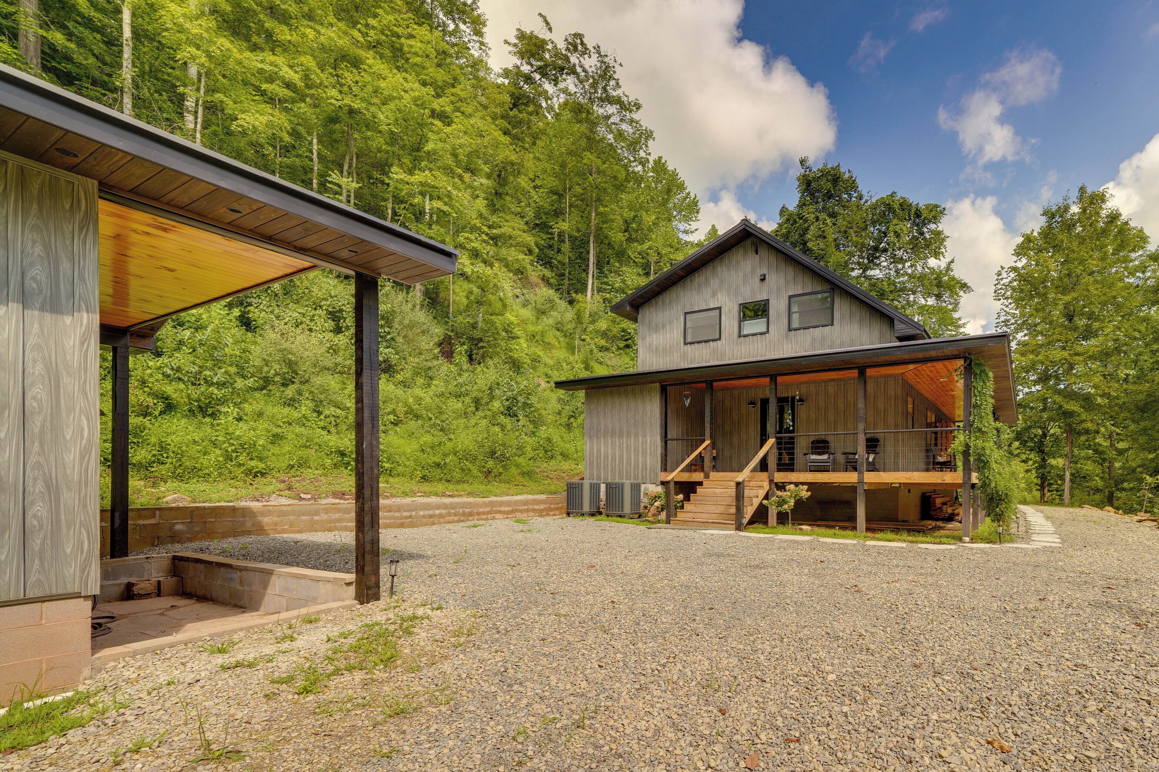 A modern wooden cabin with a covered porch and a deck, surrounded by a forested area.